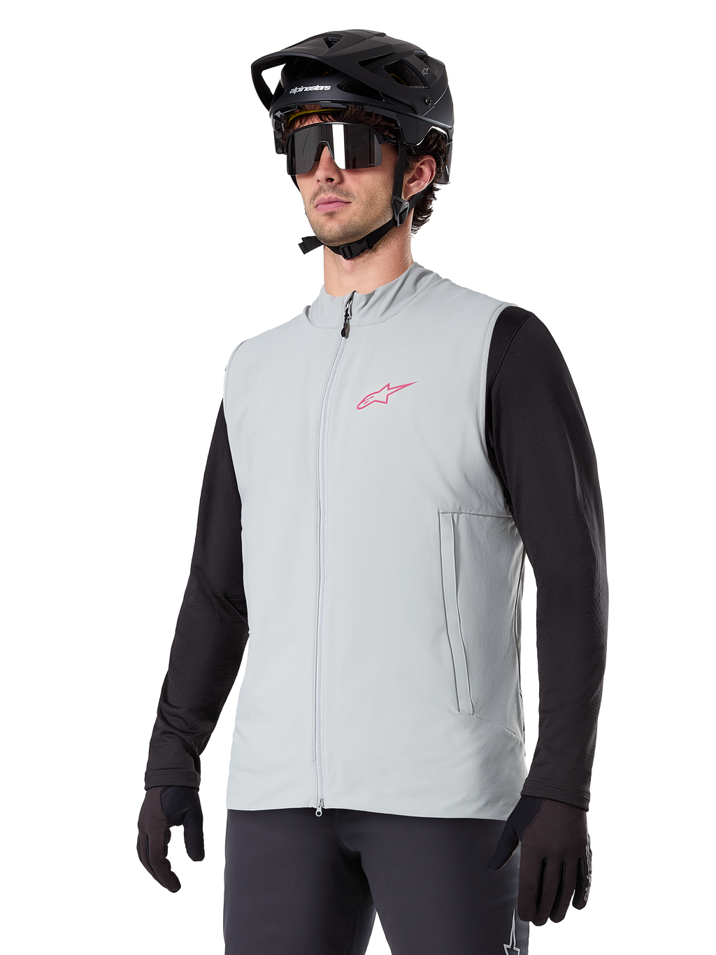Alpinestars A-Dura Thermoweste, Titanium-Grau, isolierte Fahrradweste, getragen über einem schwarzen Langarmshirt mit schwarzem Mountainbike-Helm und Handschuhen, mit pinkfarbenem Logo auf der Brust und vertikalen Reißverschlusstaschen
