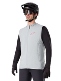 Alpinestars A-Dura Thermoweste, Titanium-Grau, isolierte Fahrradweste, getragen über einem schwarzen Langarmshirt mit schwarzem Mountainbike-Helm und Handschuhen, mit pinkfarbenem Logo auf der Brust und vertikalen Reißverschlusstaschen