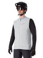 Alpinestars A-Dura Thermoweste, Titanium-Grau, isolierte Fahrradweste, getragen über einem schwarzen Langarmshirt mit schwarzem Mountainbike-Helm und Handschuhen, mit pinkfarbenem Logo auf der Brust und vertikalen Reißverschlusstaschen