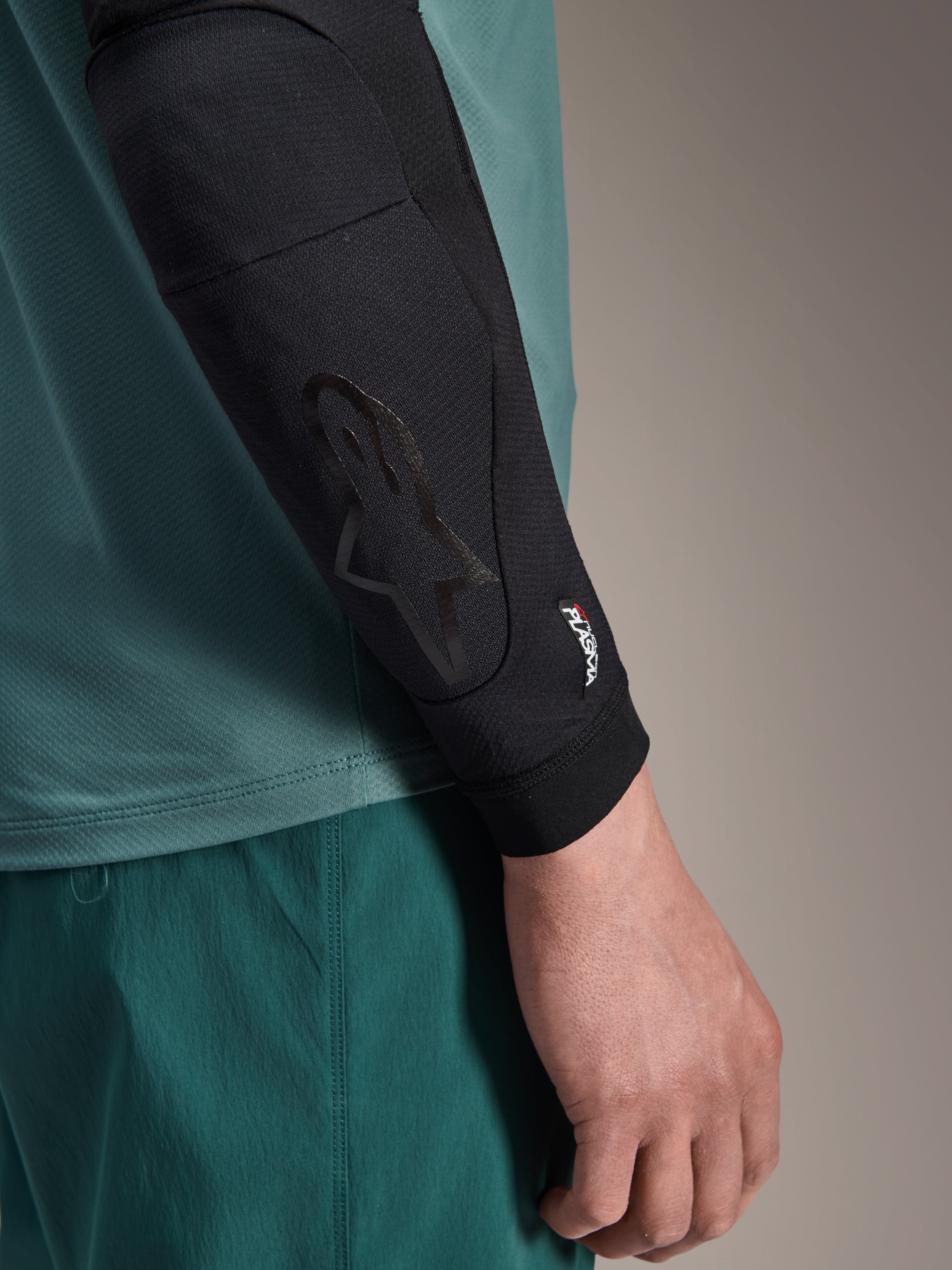 Alpinestars A-Motion Plasma Ellbogenschoner, Schwarz, MTB-Ellbogenprotektor, Nahaufnahme am Arm getragen mit petrolfarbenem Trikot, mit dezentem Astars-Logo und Nucleon Plasma Branding, schlankes Mesh-Design für den Offroad-Radsport