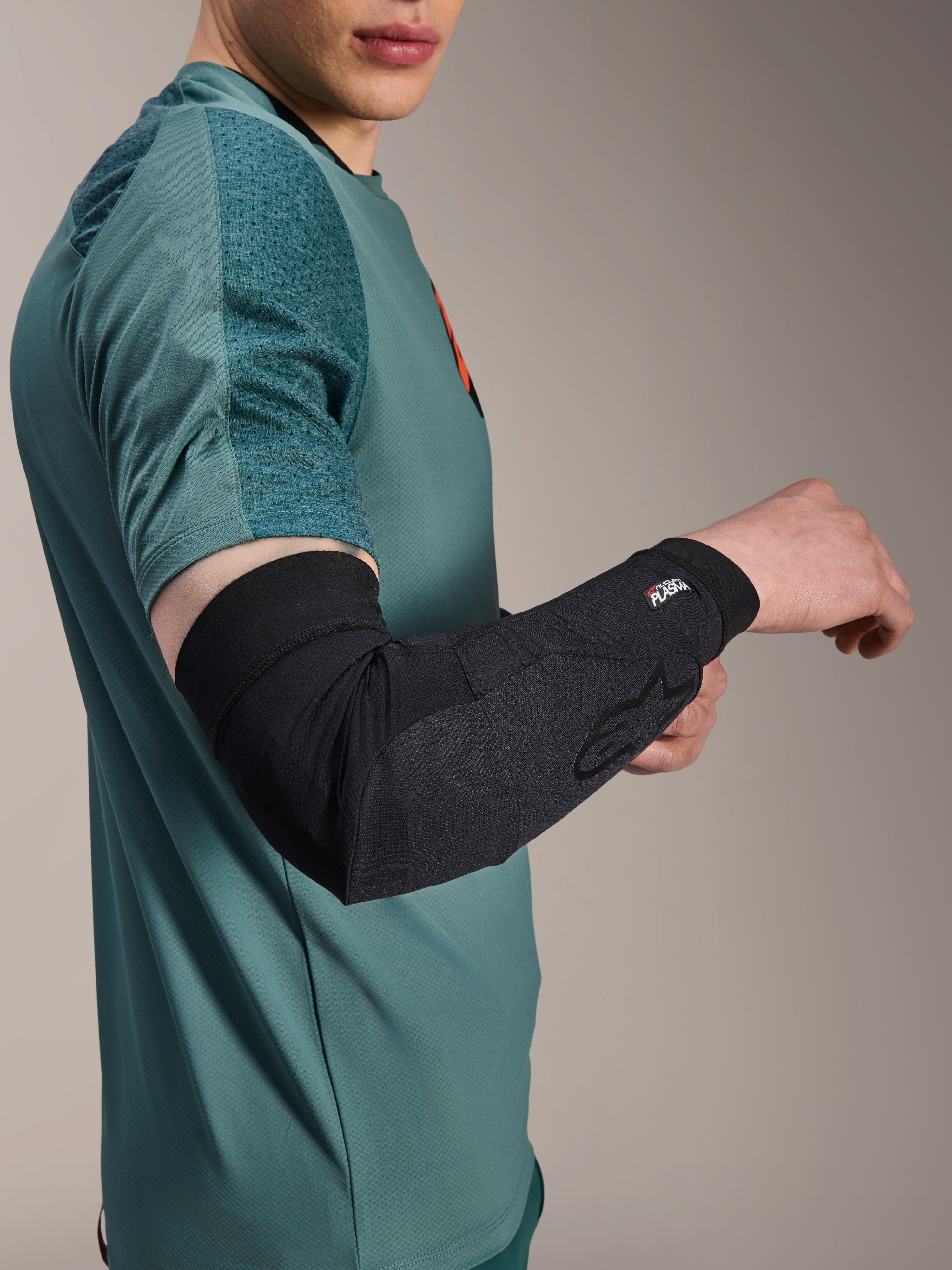 Alpinestars A-Motion Plasma Ellbogenärmel, Radsport-Ellbogenprotektor, Schwarz, getragen von einem Fahrer mit einem blaugrünen Trikot, mit Alpinestars-Logo und Nucleon Plasma-Branding, schlankes und atmungsaktives Design für MTB-Schutz