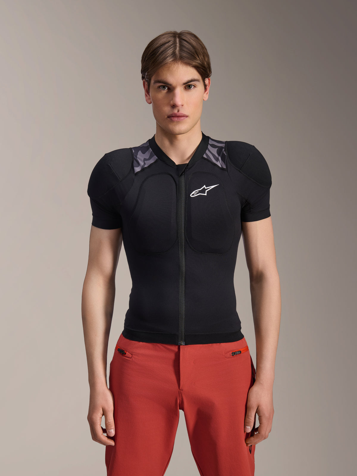 Männliches Model in Alpinestars A-Motion Plasma Schutzjacke – Kurzarm, Schwarz mit grauer Camouflage-Schulterpolsterung, weißes Alpinestars-Logo auf der Brust, kombiniert mit einer roten Hose, Slim-Fit MTB-Protektorenjacke mit Frontreißverschluss