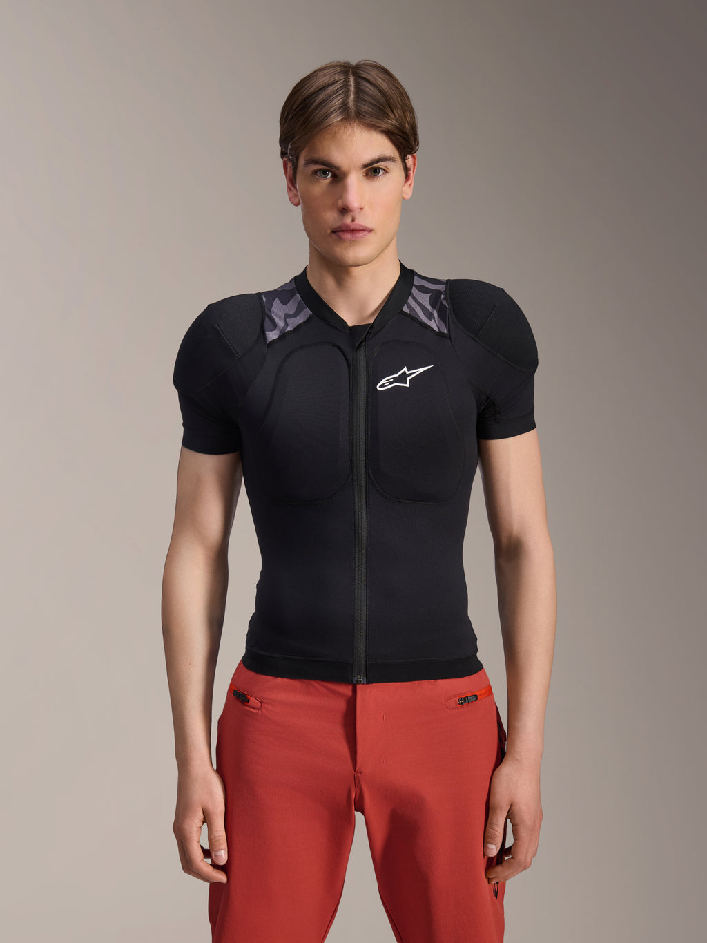 Male model wearing Alpinestars A-Motion Plasma Protection Jacket - Short Sleeve, Black with gray camo shoulder padding, white Alpinestars logo on the chest, paired with red pants, slim-fit MTB body armor with a front zipper