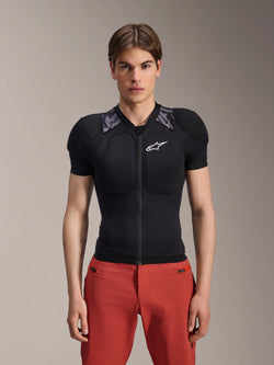 Male model wearing Alpinestars A-Motion Plasma Protection Jacket - Short Sleeve, Black with gray camo shoulder padding, white Alpinestars logo on the chest, paired with red pants, slim-fit MTB body armor with a front zipper
