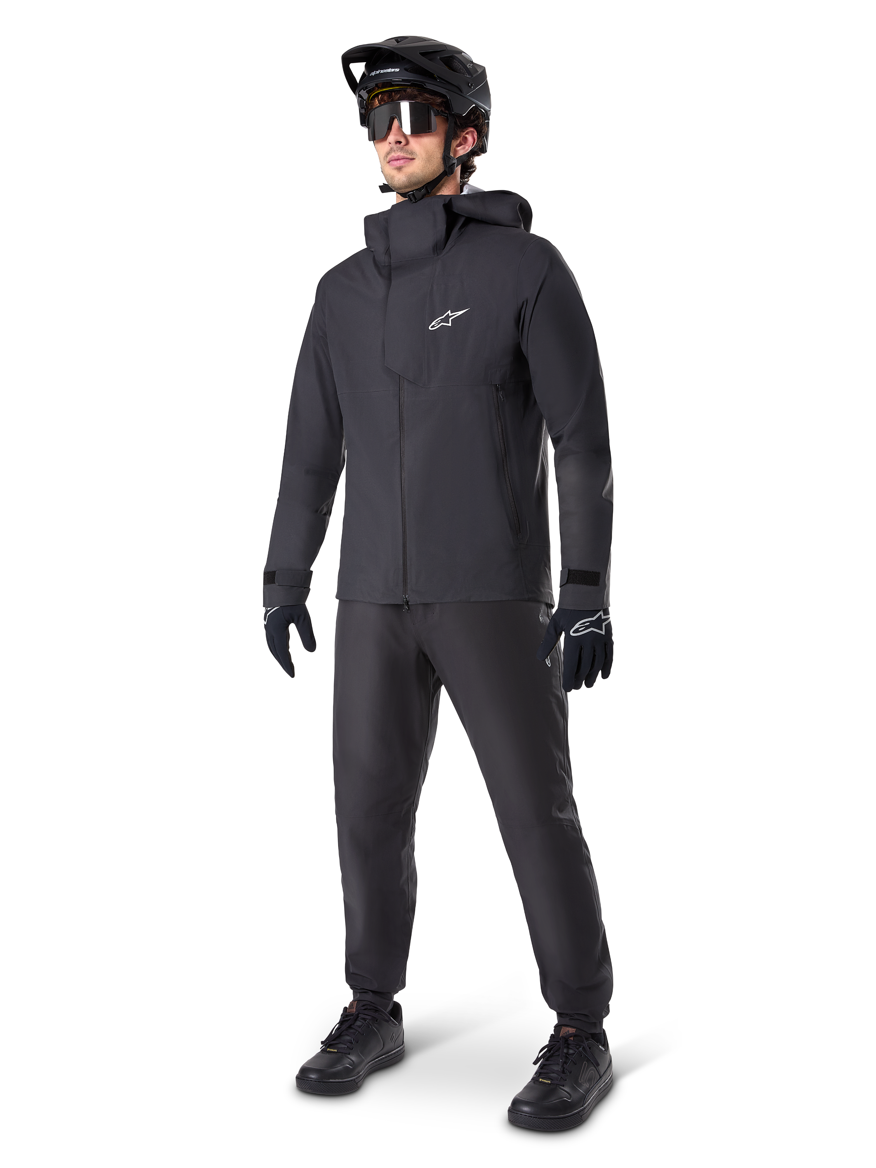 Male mountain biker wearing Alpinestars A-Dura Waterproof Pants in black, paired with a matching black hooded waterproof jacket, black MTB helmet with visor, dark cycling glasses, and black gloves, full professional gear set for all-weather trail riding