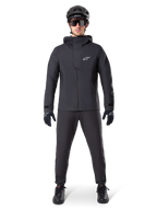Alpinestars A-Dura Waterproof Pants, black MTB pants, worn as part of a complete all-black mountain biking gear set including a technical waterproof jacket, black helmet, sunglasses, and gloves, designed for wet-weather off-road riding.