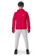 Alpinestars A-Dura Wasserdichte Hose, MTB-Hose, Titanium-Grau, getragen mit einer leuchtend roten Kapuzenjacke, roten Handschuhen und einem schwarzen Mountainbike-Helm, professionelle wasserdichte Fahrradausrüstung für nasse Trail-Bedingungen