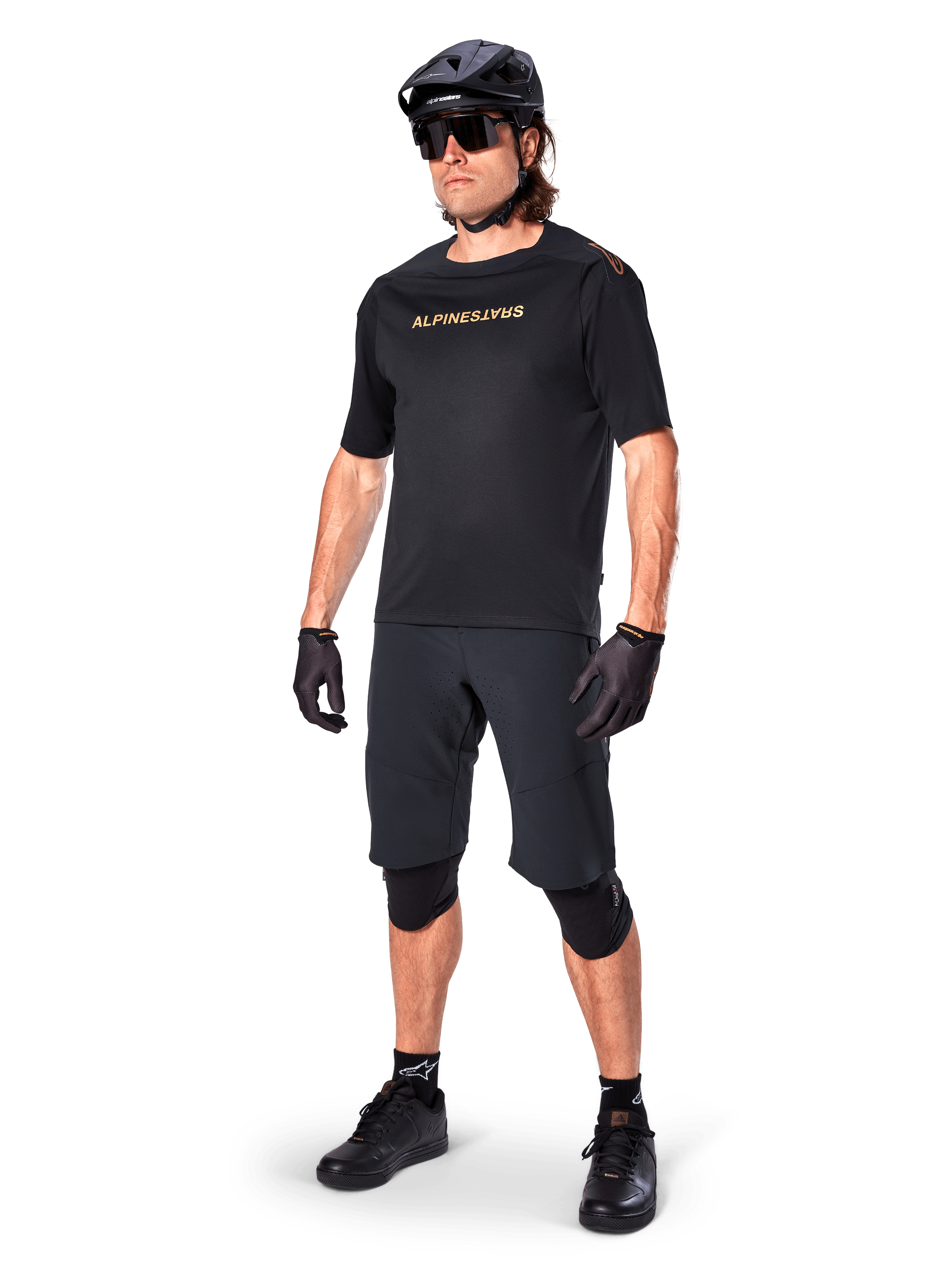 Cyclist wearing Alpinestars A-Aria Elite Shorts, Black Mountain Bike Shorts, paired with a matching black jersey with gold branding, black helmet, sunglasses, and knee pads, complete performance outfit for off-road cycling