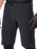 Alpinestars A-Aria Elite Shorts in Black, close-up view of the waist and thigh area, featuring magnetic buckle closure, zippered pockets, and laser-cut ventilation perforations, worn with black cycling gloves for mountain biking.