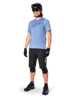 Male rider wearing Alpinestars A-Dura Elite Shorts in Black, light blue short-sleeve jersey with chest logo, black helmet, sunglasses, and protective knee pads, full mountain biking gear setup for off-road trails.