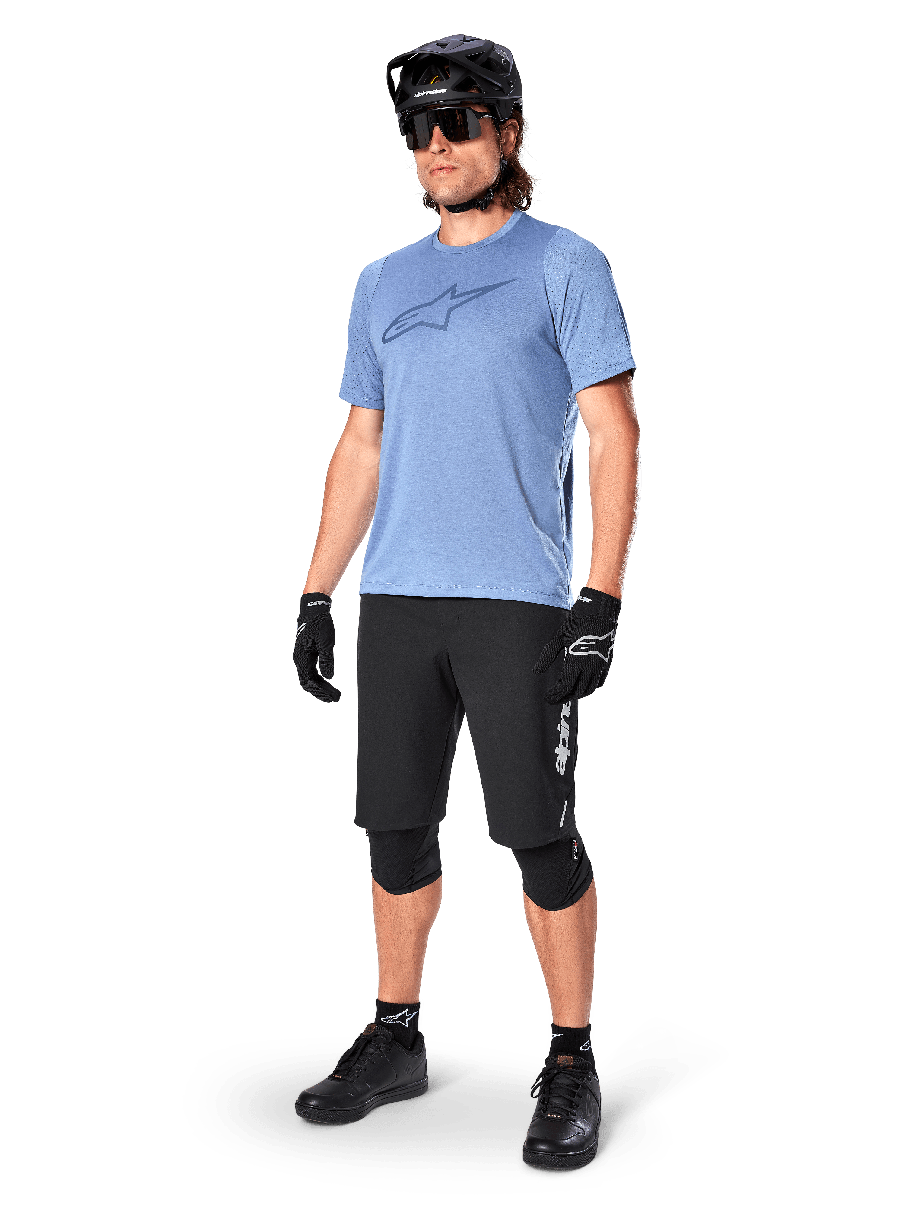 Male rider wearing Alpinestars A-Dura Elite Shorts in Black, light blue short-sleeve jersey with chest logo, black helmet, sunglasses, and protective knee pads, full mountain biking gear setup for off-road trails.