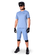 Model in Alpinestars A-Dura Shorts in Infinity Blue, passendes hellblaues Kurzarm-Trikot, schwarzer MTB-Helm, Sonnenbrille und schwarze Handschuhe, komplettes Mountainbike-Outfit für Trailfahrten