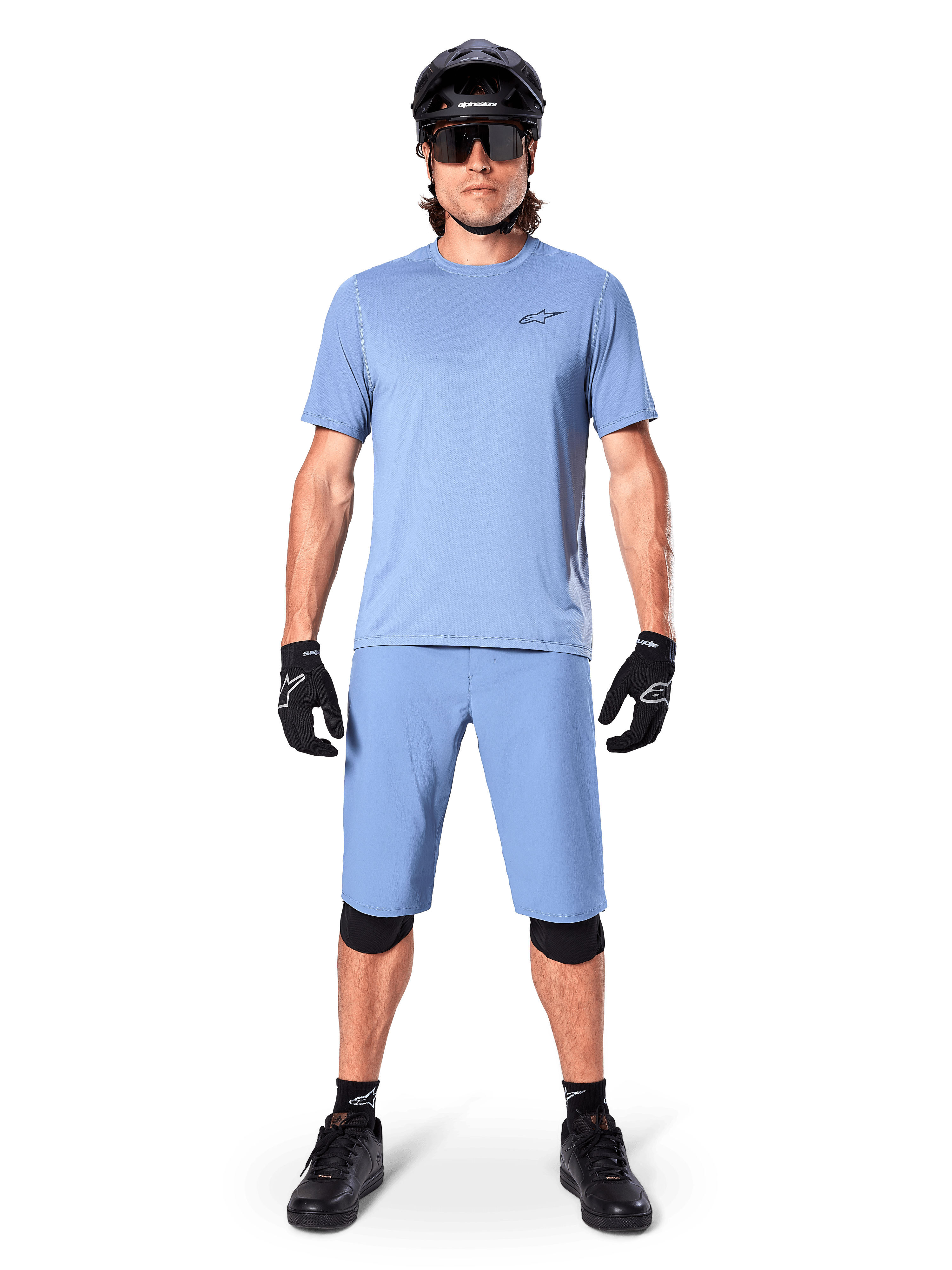 Model trägt Alpinestars A-Dura Shorts in Infinity Blue, passendes hellblaues Trikot, schwarzen Mountainbike-Helm, schwarze Sonnenbrille, schwarze Handschuhe und Knieschoner, komplettes MTB-Outfit für Trailfahrten