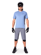 Male rider wearing Alpinestars A-Dura Shorts in Dark Gray, styled with a light blue short-sleeve t-shirt, black helmet, sunglasses, and black knee pads, complete mountain biking outfit for off-road performance.
