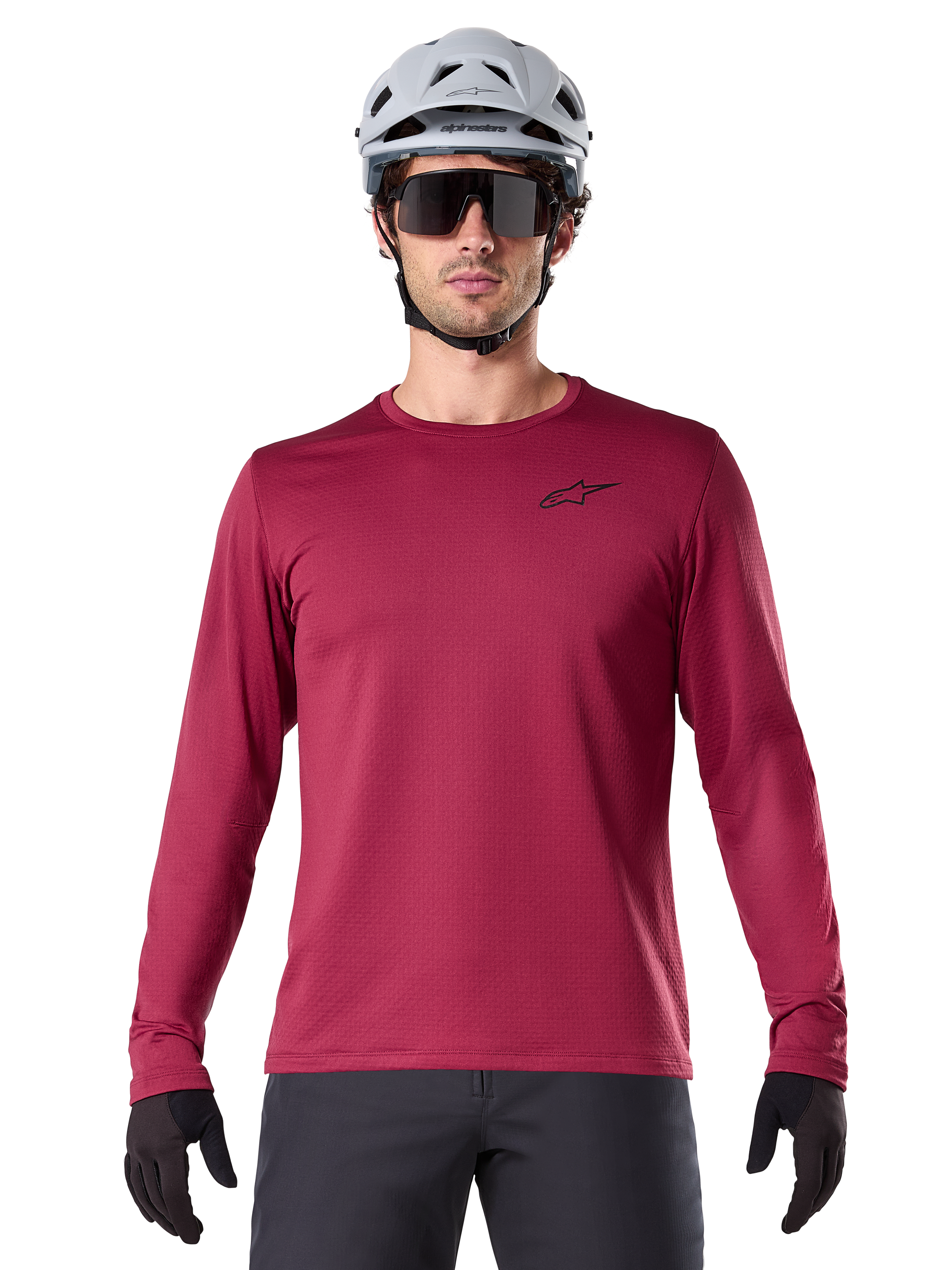 Männlicher Radfahrer trägt das Alpinestars A-Dura Thermal Jersey, langärmeliges MTB-Trikot, Burgunderrot, kombiniert mit einem grauen Helm und einer schwarzen Sonnenbrille, kleines schwarzes Stern-Logo auf der Brust, isoliertes Fleece-Material für Mountainbiking bei kaltem Wetter