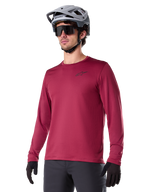 Alpinestars A-Dura Thermal Jersey, Langarm-Mountainbike-Trikot, Burgunderrot, mit isoliertem Fleece-Futter und kleinem schwarzem Brust-Logo, getragen mit einem grauen Helm, schwarzer Sonnenbrille und schwarzen Handschuhen, ideal für Radsport bei kaltem Wetter