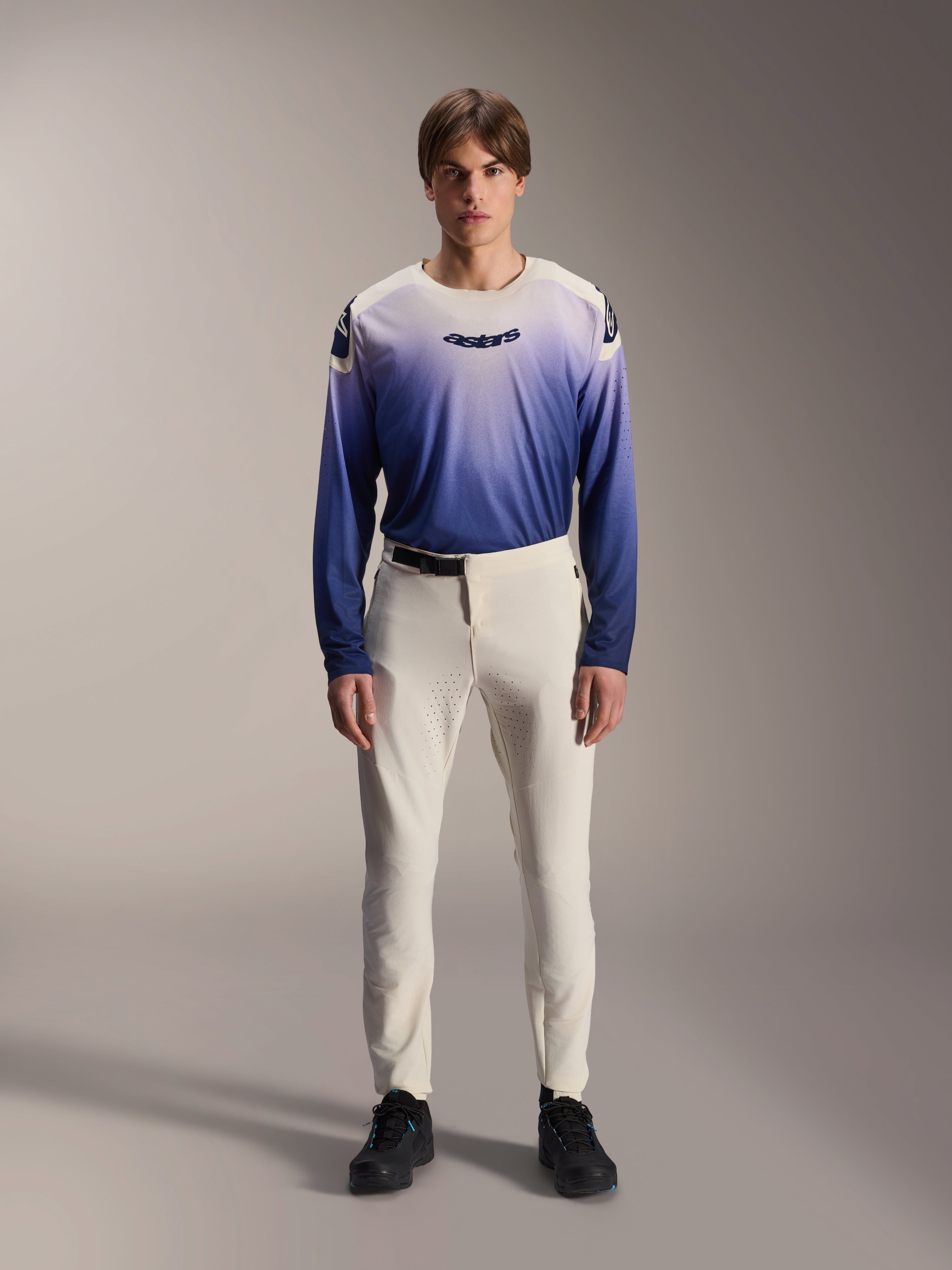 Alpinestars A-Aria Elite Hose, Ecru Performance MTB-Hose, naturweiße Radhose mit laserperforierten Oberschenkel-Panels und Magnetschnalle, getragen mit blauem Langarmtrikot mit Farbverlauf und schwarzen Schuhen, Performance-Passform für Trailriding
