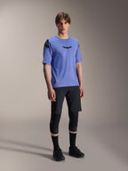 Männliches Modell in Alpinestars A-Aria Elite Shorts in Black Titanium und A-Aria Polartec Elite SS Jersey in Helllila, kombiniert mit schwarzen Mountainbike-Knieschützern und Schuhen, professionelle Radsportausrüstung für Offroad-Performance