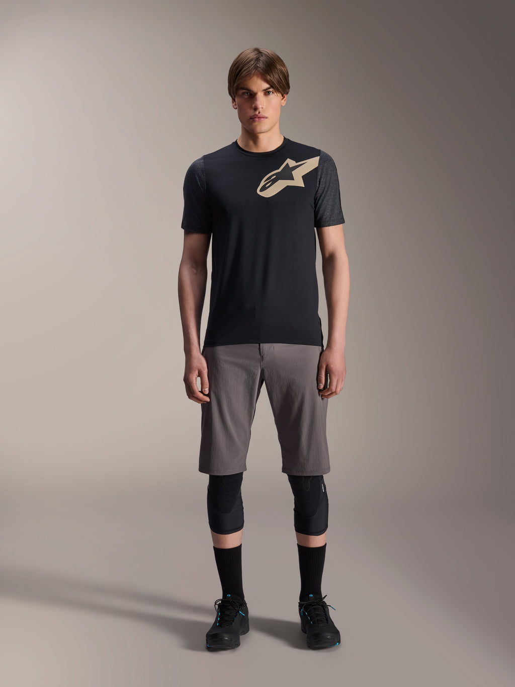 Alpinestars A-Dura Lipan Kurzarmtrikot, Schwarz mit grau melierten Ärmeln und beigem Astars-Logo auf der Brust, getragen von einem männlichen Model mit grauen Mountainbike-Shorts und schwarzen Knieprotektoren, Performance-MTB-Ausrüstung