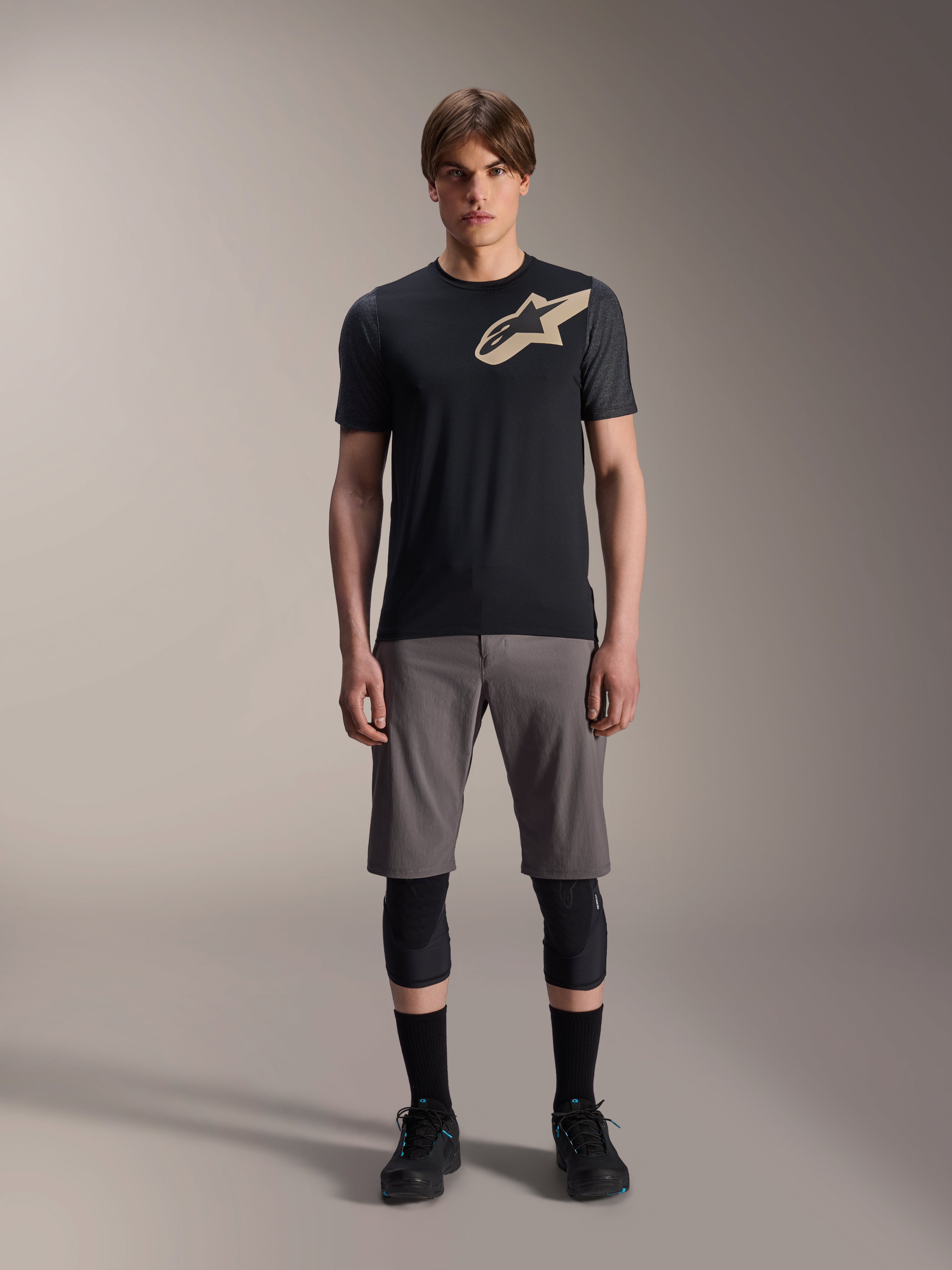 Alpinestars A-Dura Lipan Kurzarmtrikot, Schwarz mit grau melierten Ärmeln und beigem Astars-Logo auf der Brust, getragen von einem männlichen Model mit grauen Mountainbike-Shorts und schwarzen Knieprotektoren, Performance-MTB-Ausrüstung