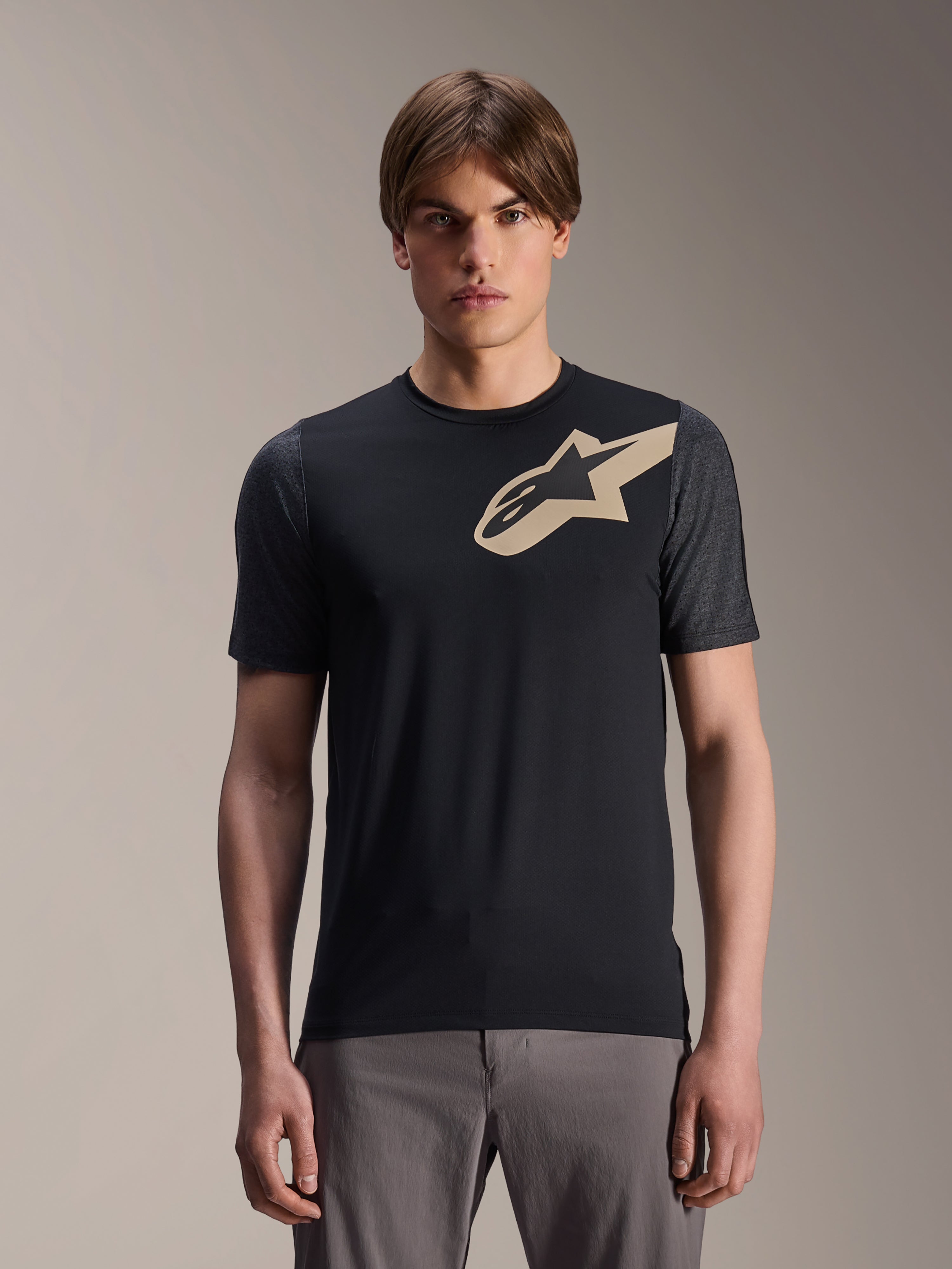 Männliches Model im Alpinestars A-Dura Lipan Kurzarmtrikot, schwarzes MTB-Mountainbike-Trikot mit beigem Stern-Logo und dunkelgrauen Ärmeln, graue Radhose, lässige Performance-Bekleidung