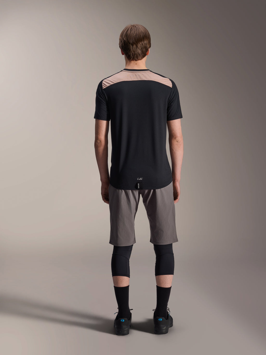 Alpinestars A-Dura Lipan Kurzarmtrikot, Schwarz mit hellbraunem Einsatz am oberen Rücken, Rückansicht mit verlängertem Rückensaum, getragen mit grauen MTB-Shorts und schwarzen Knieprotektoren, Performance-Mountainbike-Bekleidung