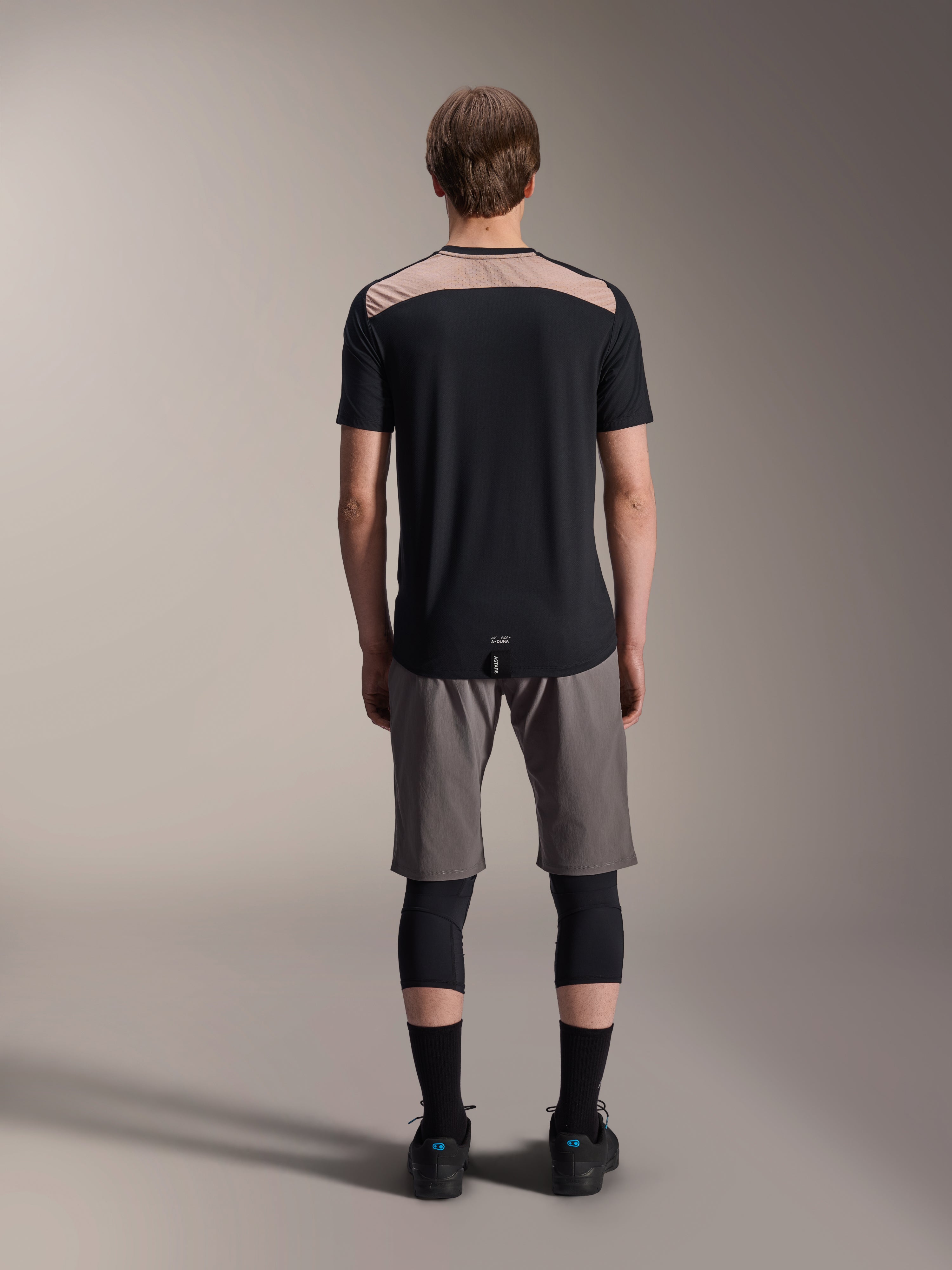 Alpinestars A-Dura Lipan Kurzarmtrikot, Schwarz mit hellbraunem Einsatz am oberen Rücken, Rückansicht mit verlängertem Rückensaum, getragen mit grauen MTB-Shorts und schwarzen Knieprotektoren, Performance-Mountainbike-Bekleidung