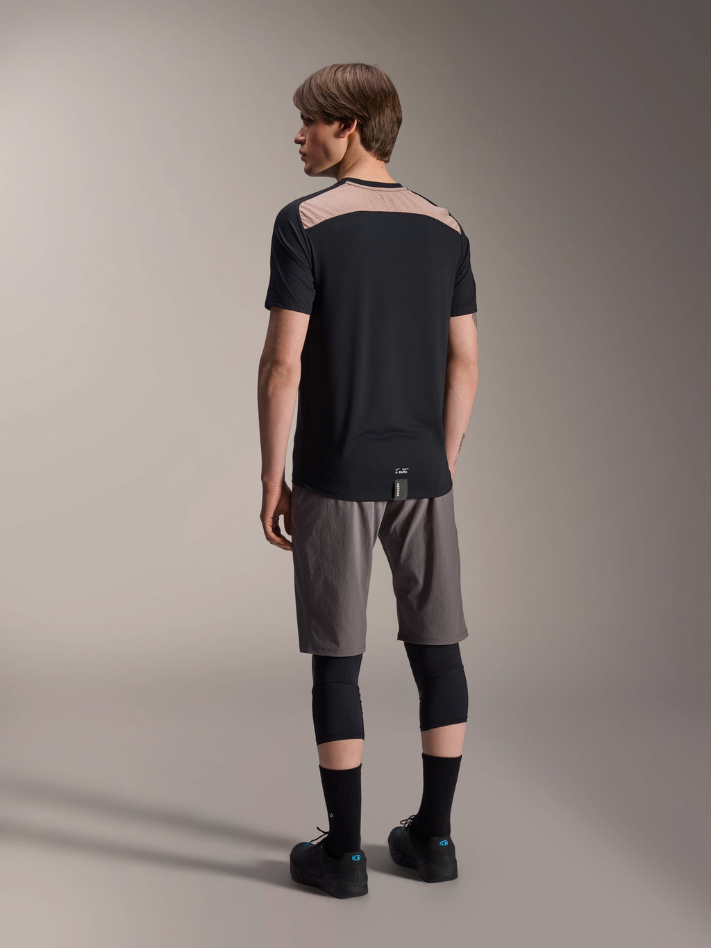 Alpinestars A-Dura Lipan Kurzarmtrikot in Schwarz, Rückansicht am Model zeigt den hellbraunen perforierten Mesh-Einsatz am oberen Rücken, kombiniert mit grauen Mountainbike-Shorts und schwarzen Knieprotektoren für ein komplettes Trail-Outfit.
