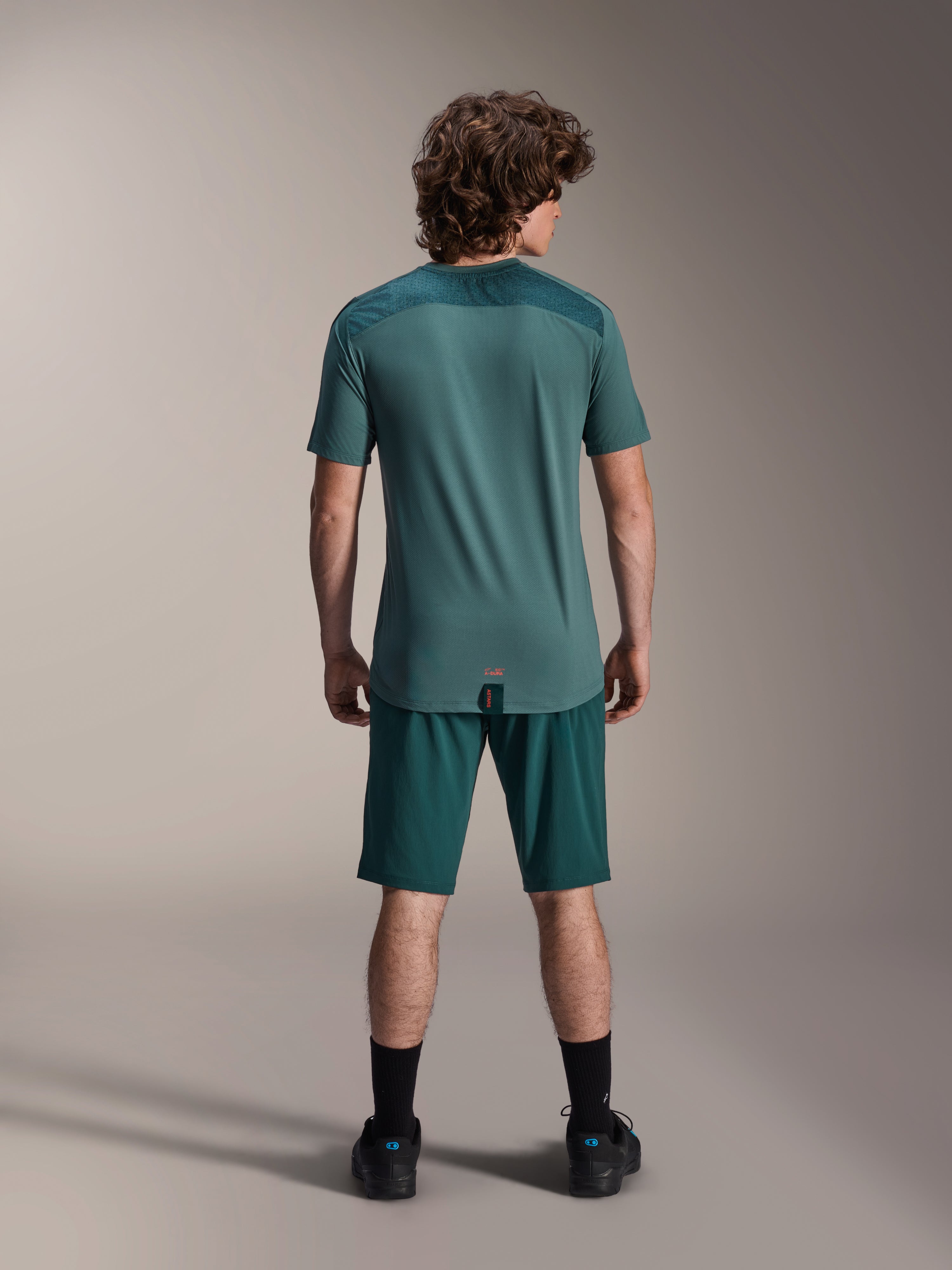 Alpinestars A-Dura Lipan Short Sleeve Jersey in Silver Pine, back view showing the textured jacquard shoulder panels and drop tail hem, worn with matching dark green mountain bike shorts and black footwear.