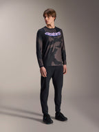 Männliches Modell im Alpinestars A-Dura Hollow Camo Langarm-Trikot, Black Camo Muster mit lilafarbener 'astars' Brustgrafik, kombiniert mit schwarzer technischer Hose und schwarzen Schuhen, lässige MTB-Ausrüstung