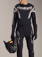 Alpinestars A-Supra Melt Long Sleeve Jersey, Black with grey graphic panels and bold 'astars' wordmark on chest, worn with black mountain bike pants and gloves while holding a black full-face helmet, performance MTB racing kit