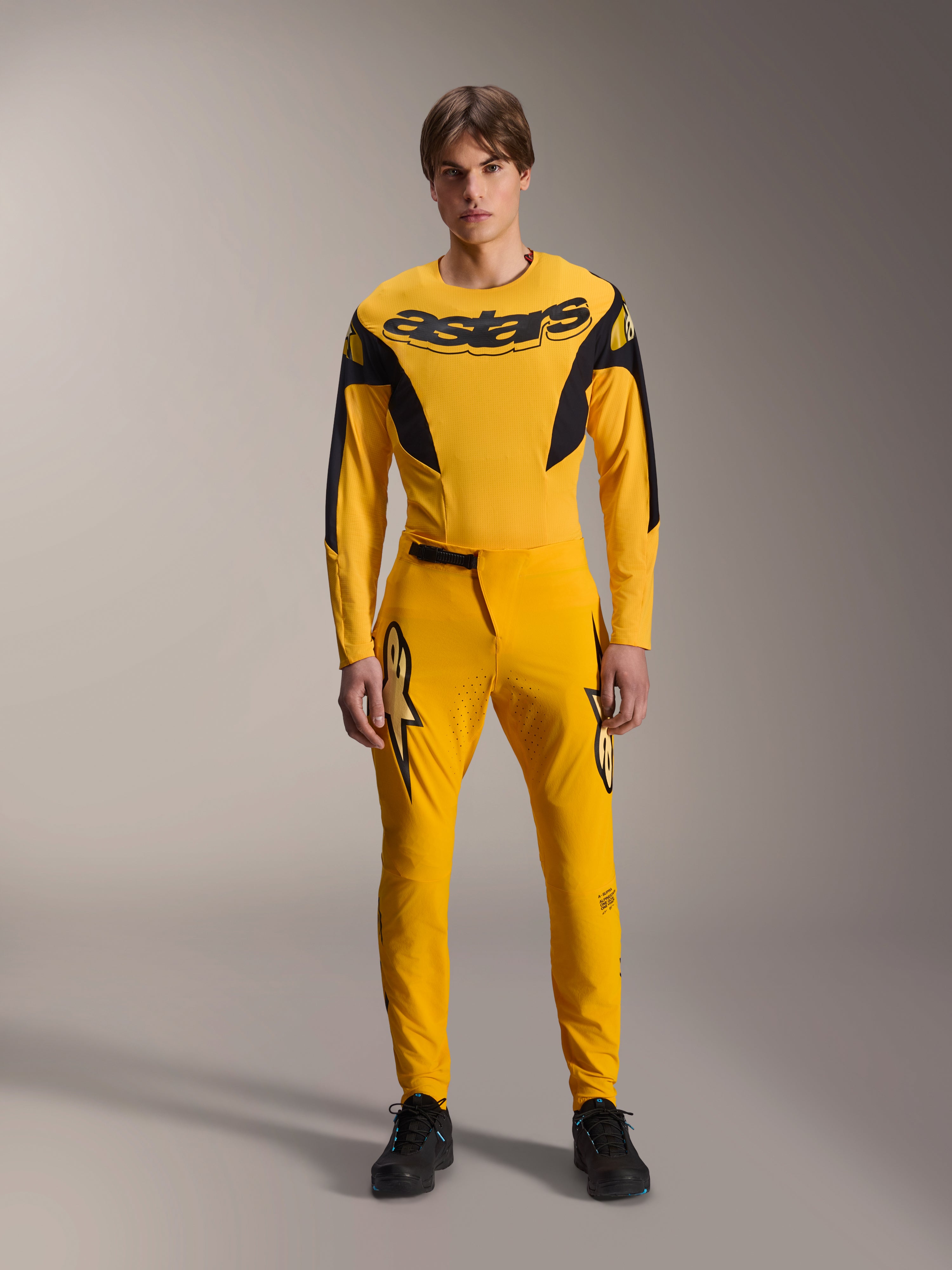 Limited Edition A-Supra Melt Gold Gear-Set, Alpinestars Mountainbike-Bekleidung, Dunkelgelb und Schwarz, Modell trägt langärmeliges Trikot mit markantem 'astars'-Brustlogo und passender Performance-Hose, lasergeschnittene Belüftung an den Innenseiten der Oberschenkel, Ratschenverschluss, professionelle MTB-Rennausrüstung