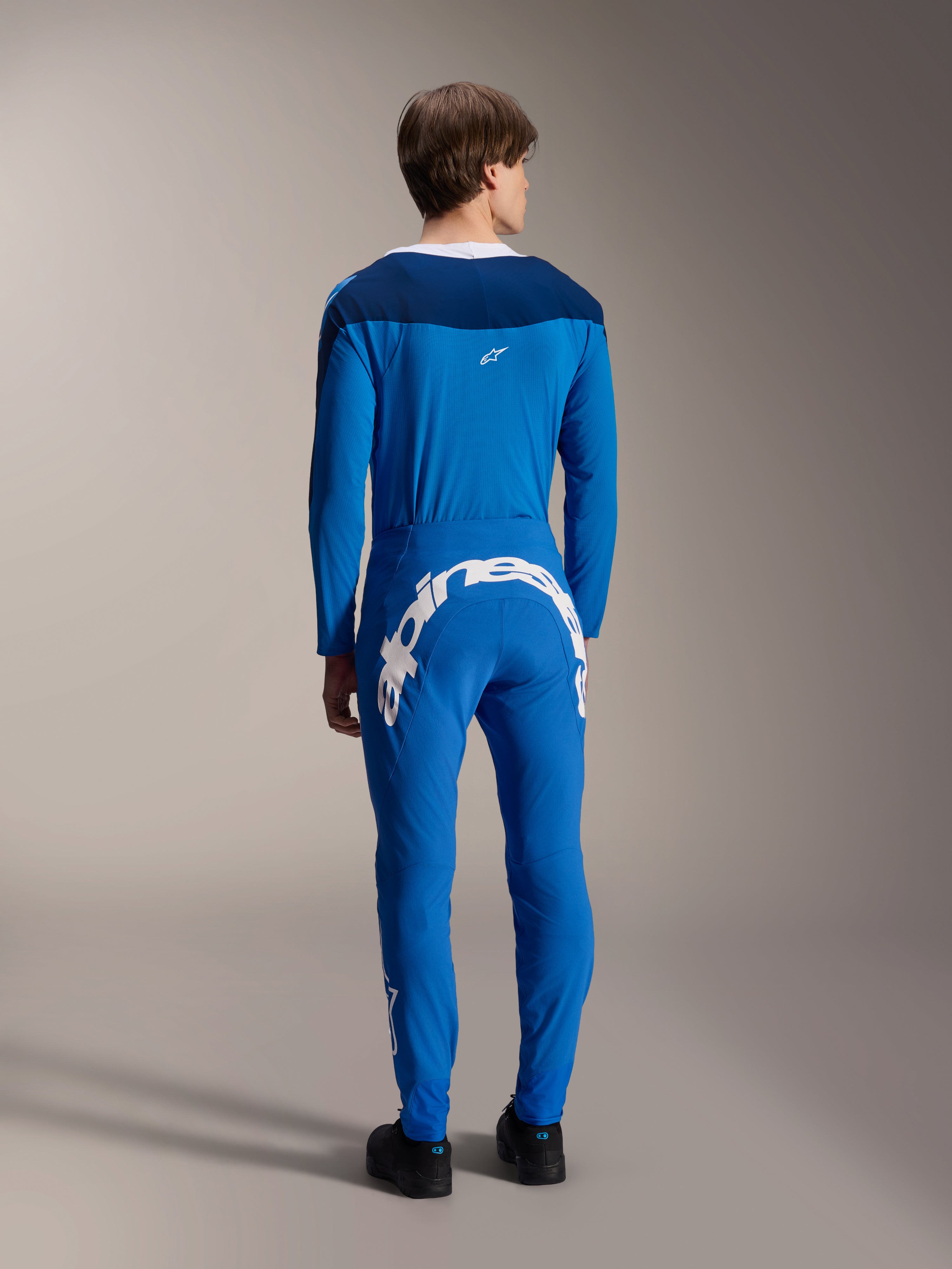 Männliches Modell von hinten gesehen im Alpinestars A-Supra Melt Langarm-Trikot in Blau, kombiniert mit passender blauer Performance-Hose mit großem weißem Alpinestars-Schriftzug auf der Rückseite, komplette MTB-Rennausrüstung.