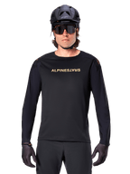 Alpinestars A-Aria Polartec Switch Long Sleeve Jersey, Black, gold Alpinestars logo on chest, worn with a black MTB helmet, dark sunglasses, and black gloves, professional mountain bike trail gear