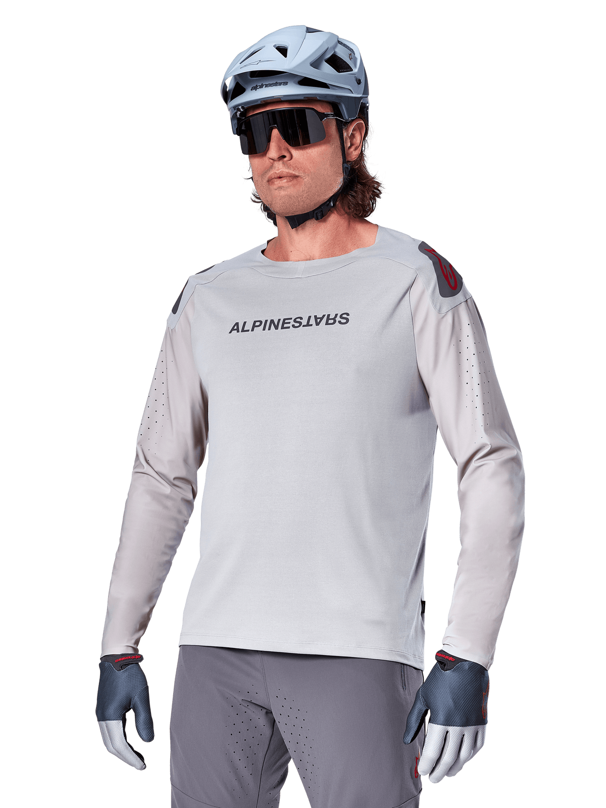 Alpinestars A-Aria Polartec Switch Langarmtrikot, hellgraues Radtrikot mit schwarzem Brustlogo und roten Schulterakzenten, laserperforierte Ärmel für optimale Luftzirkulation, getragen mit hellblauem Helm, Sonnenbrille und grauer Hose, Performance-Mountainbike-Bekleidung für Trailfahrten