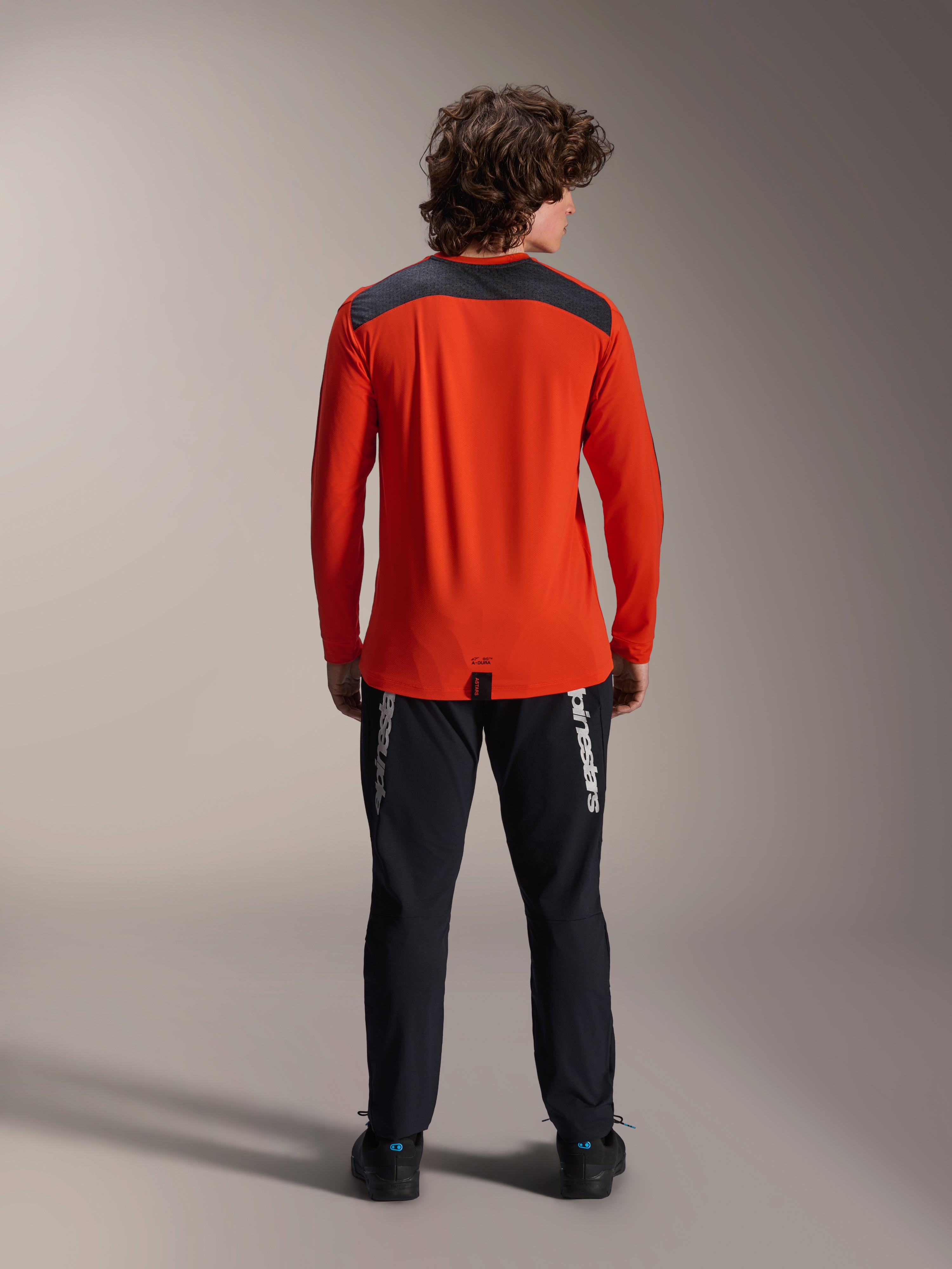 Alpinestars A-Dura Insert Astar Langarm-Trikot in Spicy Orange, Rückansicht mit anthrazitgrauem Schulterpanel, getragen mit schwarzer Alpinestars Hose und schwarzen Schuhen, professionelle Studioaufnahme für MTB-Performance-Ausrüstung