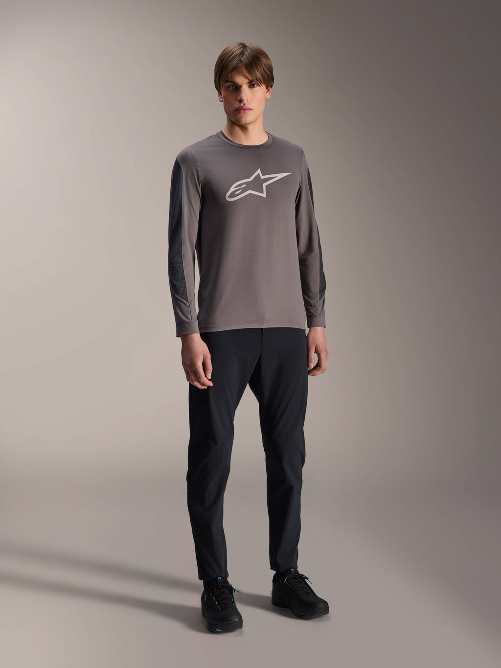 Alpinestars A-Dura Insert Astar Trikot, Langarm-MTB-Trikot, Dunkelgrau, mit großem weißem Brustlogo, getragen mit schwarzer technischer Hose und schwarzen Schuhen, Offroad-Radsportbekleidung