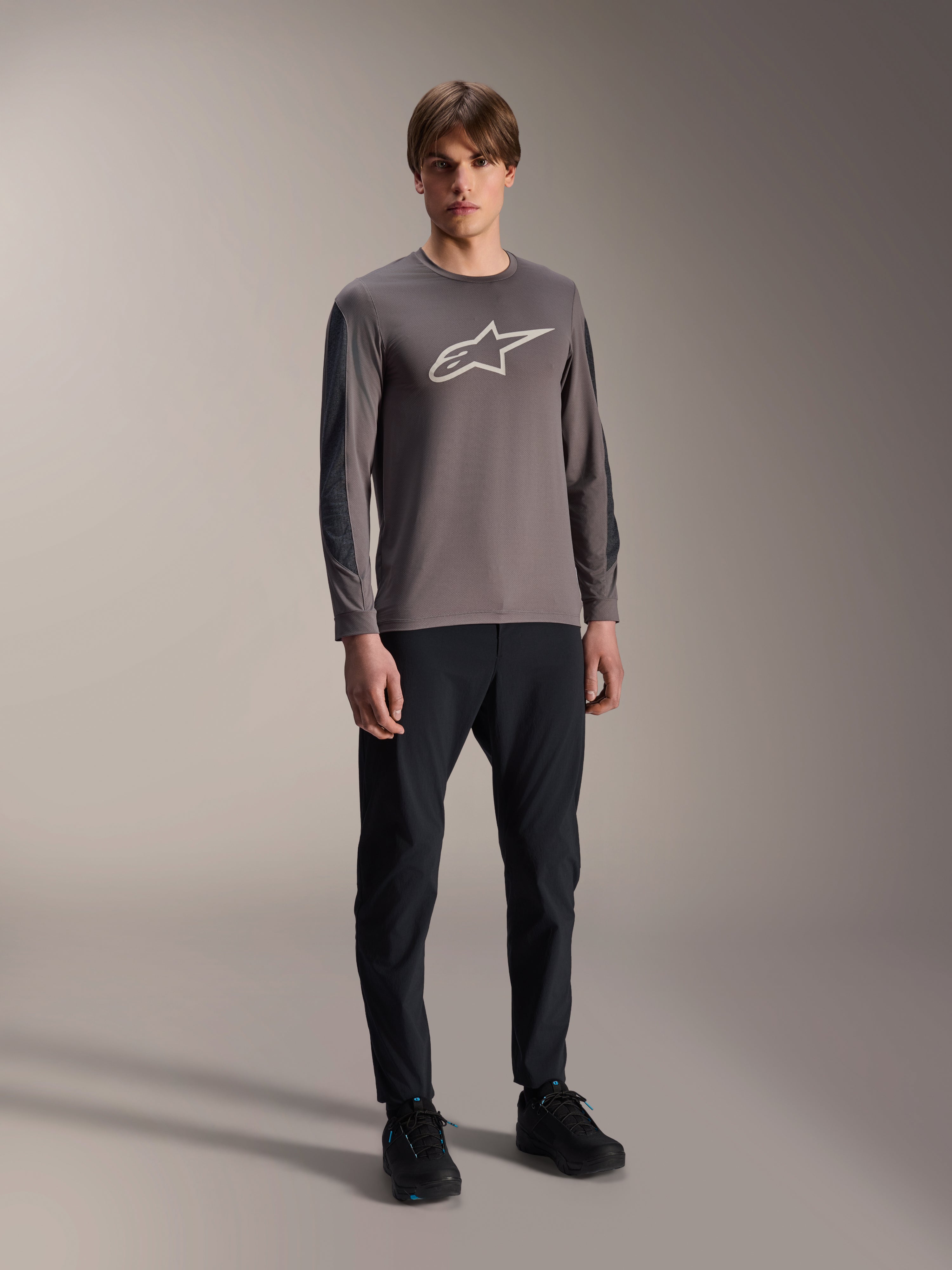 Alpinestars A-Dura Insert Astar Trikot, Langarm-MTB-Trikot, Dunkelgrau, mit großem weißem Brustlogo, getragen mit schwarzer technischer Hose und schwarzen Schuhen, Offroad-Radsportbekleidung