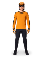 Mountain biker wearing Alpinestars A-Dura Dri Wool Long Sleeve Jersey, Dark Gold with black accents on neck and sleeves, paired with black pants, matching gold helmet, gloves, and black sunglasses, professional mountain biking gear