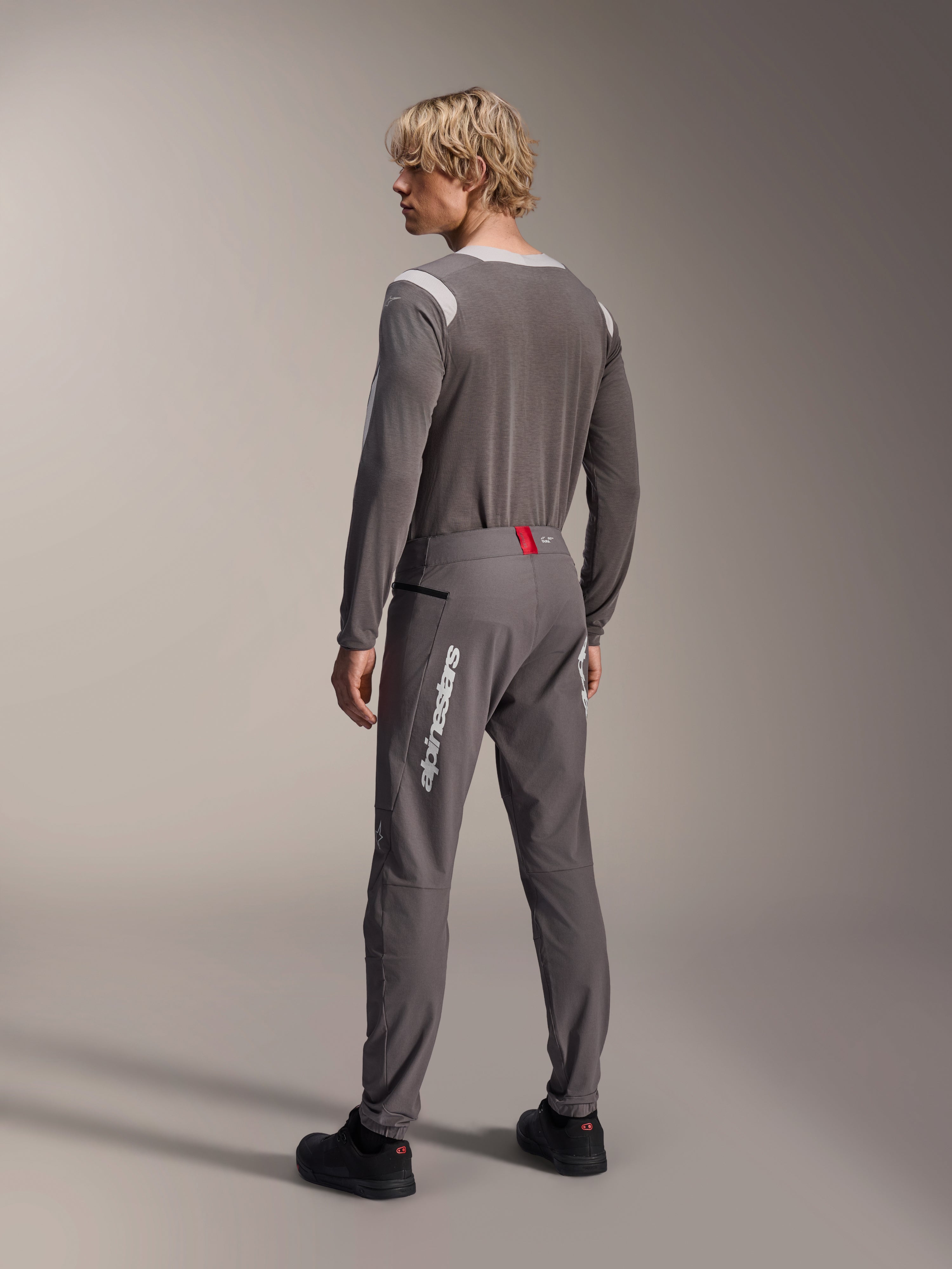 Model from a rear view wearing Alpinestars MTB gear, gray A-Dura Dri Wool Long Sleeve Jersey, Dark Gray A-Dura Elite Pants with white vertical branding and red waistband detail, coordinated cycling outfit for mountain biking