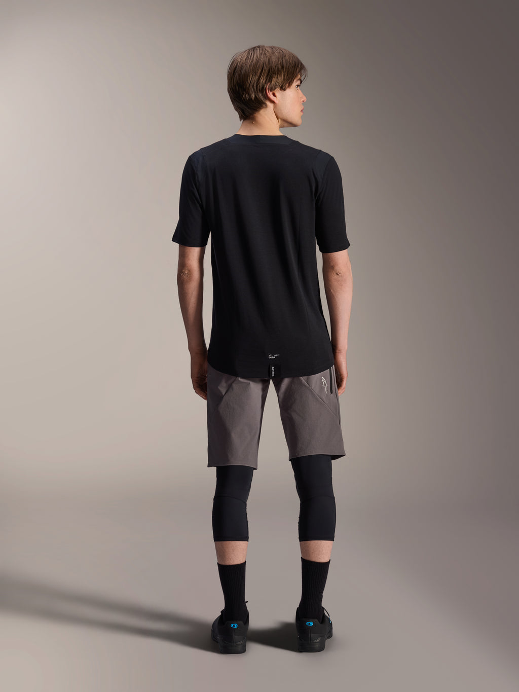 Alpinestars A-Dura Elite Shorts, Dark Gray mountain bike shorts, back view, worn with a black short-sleeve jersey, knee pads, and black socks, technical cycling gear for off-road riding