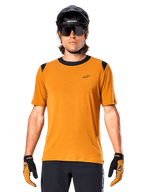 Alpinestars A-Dura Dri Wool Short Sleeve Jersey, Dark Gold, worn with black MTB helmet, sunglasses, and gloves, featuring black collar and shoulder panels, breathable drirelease wool blend for mountain biking