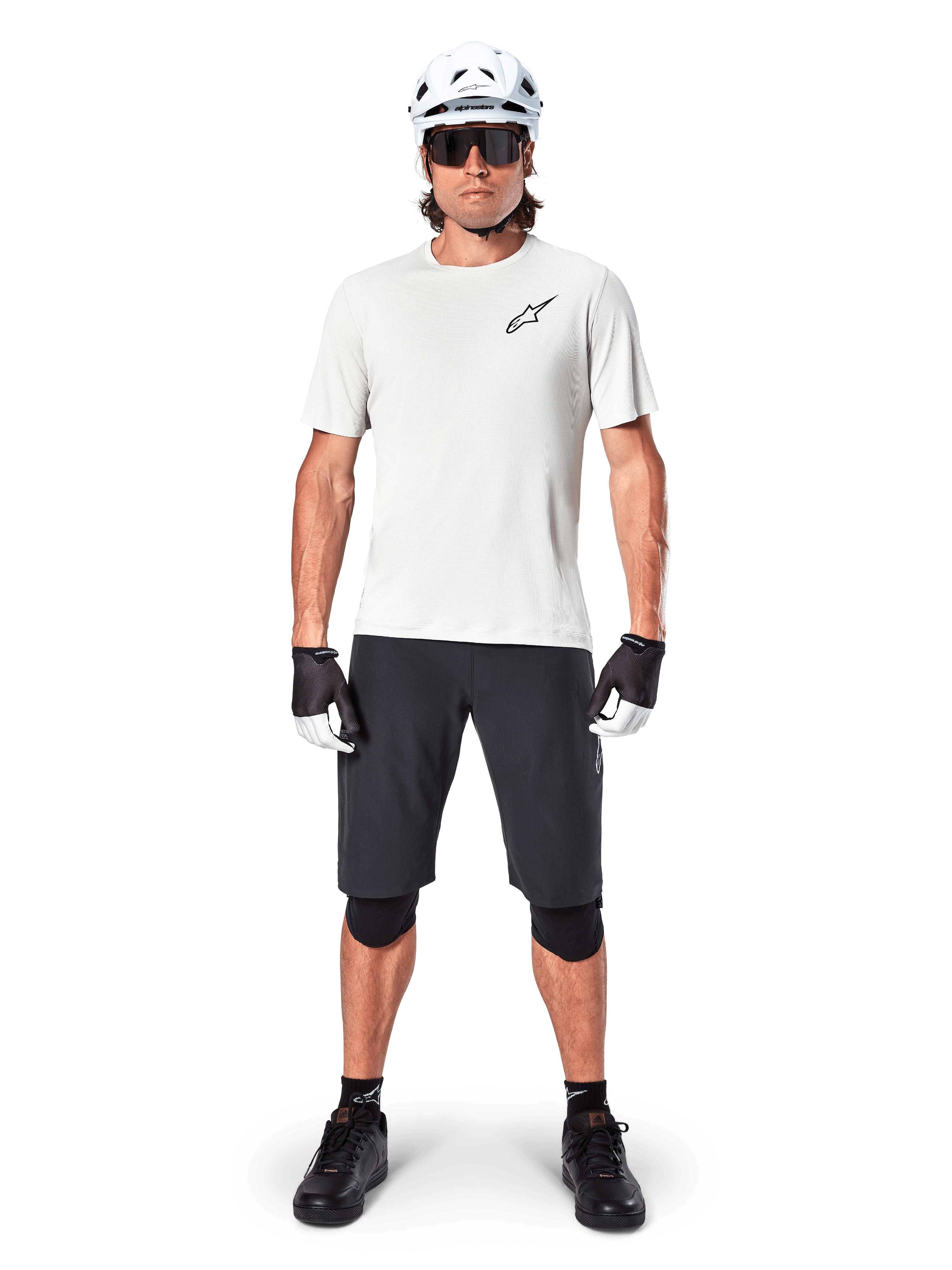 Male cyclist wearing Alpinestars A-Aria Astar Short Sleeve Jersey in White Dove, featuring a black Astars logo on the chest, paired with black mountain bike shorts, white helmet, sunglasses, black knee guards, and gloves, complete off-road cycling outfit.