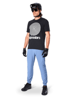 Alpinestars A-Dura Oscar Short Sleeve Jersey, Black MTB jersey featuring a large white heritage spiral graphic, worn by a rider with a black helmet, sunglasses, black gloves, and light blue trousers, performance mountain bike gear