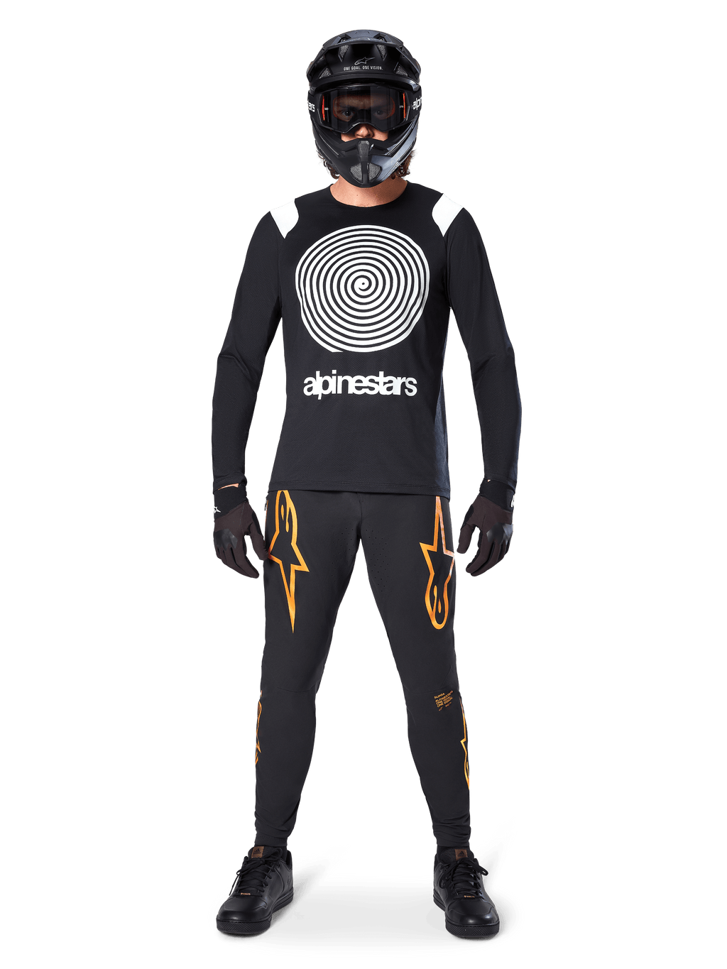 Male model wearing complete mountain bike gear, Alpinestars A-Supra Oscar Long Sleeve Jersey in black with white spiral chest graphic, black pants with orange star logos, black full-face helmet and goggles, black gloves and shoes, coordinated outfit for MTB gravity riding