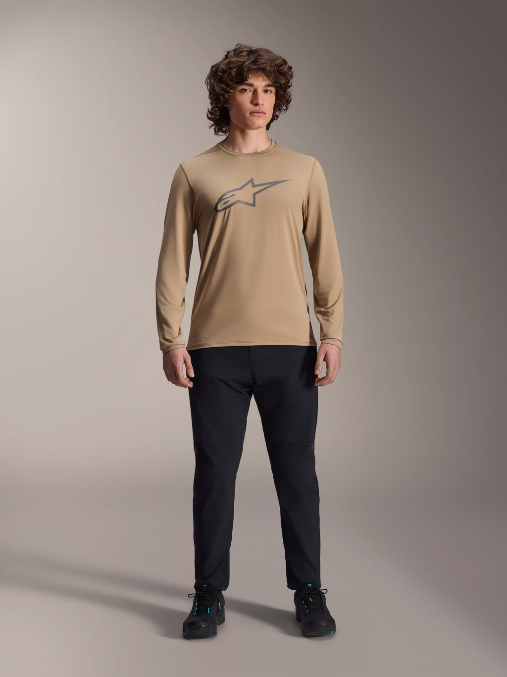 Alpinestars A-Dura Astar Langarmtrikot, Hellbraun mit grauem Astars-Brustlogo, getragen von einem Model mit schwarzer technischer Hose und Schuhen, lässige Mountainbike-Performance-Ausrüstung