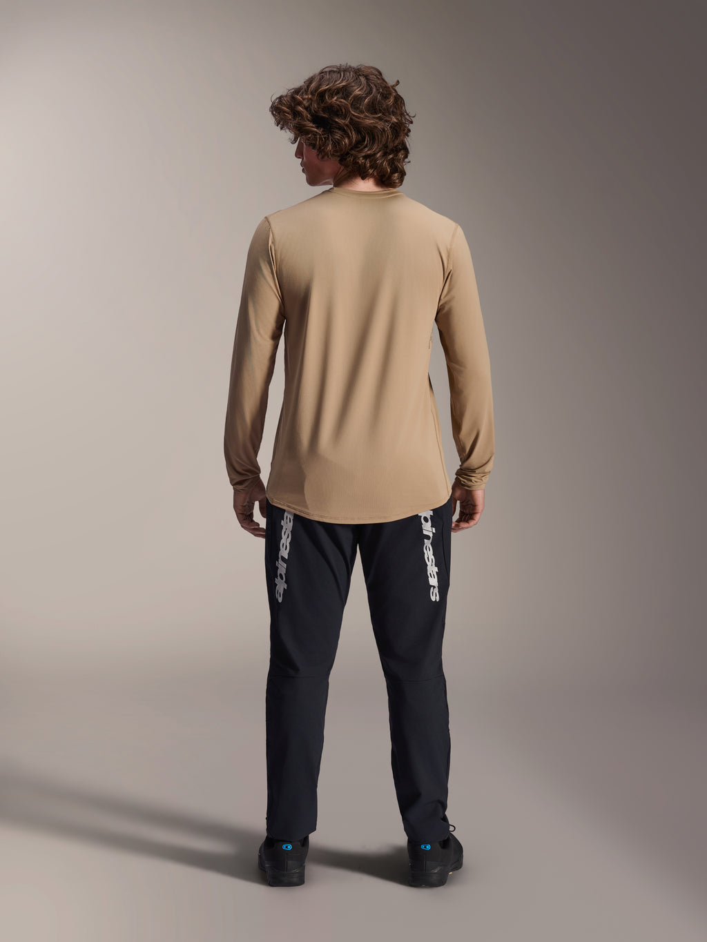 Alpinestars A-Dura Astar Langarmtrikot, Hellbraun, Rückansicht mit dem längeren abfallenden Saum, getragen mit einer schwarzen Hose mit Alpinestars-Branding, Performance-Mountainbike-Bekleidung