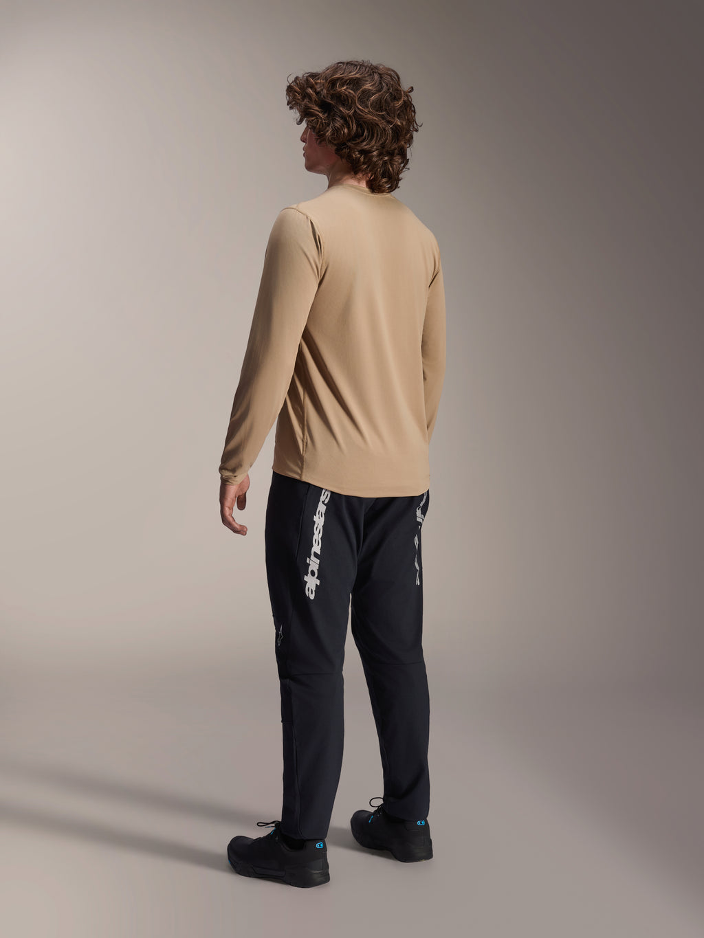 Alpinestars A-Dura Astar Langarmtrikot in Hellbraun, Rückansicht eines männlichen Models zur Präsentation des Drop-Tail-Designs, kombiniert mit schwarzer Hose und Schuhen, lässige Mountainbike-Performance-Ausrüstung.