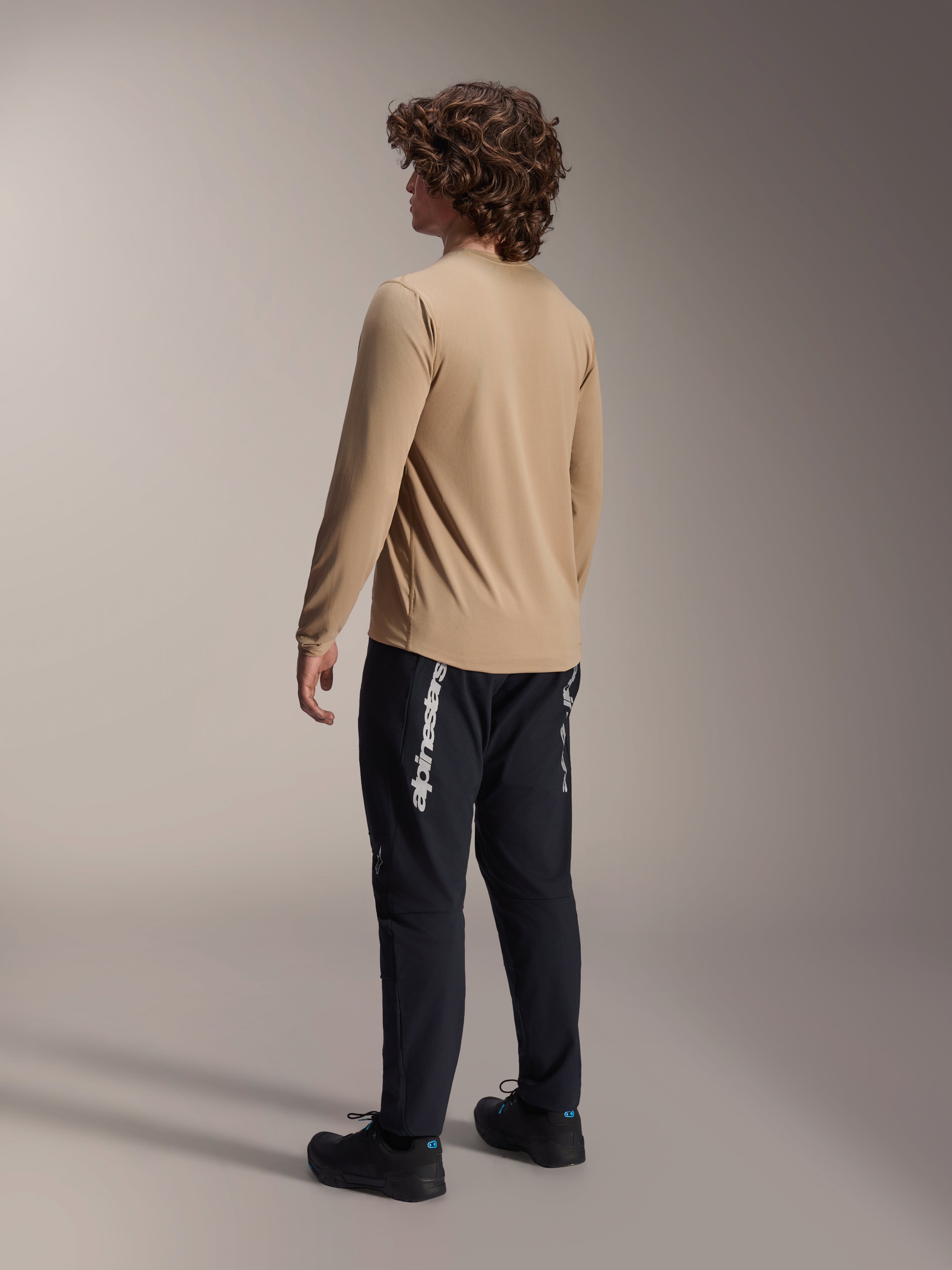 Alpinestars A-Dura Astar Langarmtrikot in Hellbraun, Rückansicht eines männlichen Models zur Präsentation des Drop-Tail-Designs, kombiniert mit schwarzer Hose und Schuhen, lässige Mountainbike-Performance-Ausrüstung.