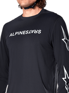 Alpinestars A-Dura Switch Long Sleeve Jersey, Black, featuring white Alpinestars wordmark on the chest and star logos on the sleeve, worn by a rider with a helmet strap visible, technical mountain bike apparel.